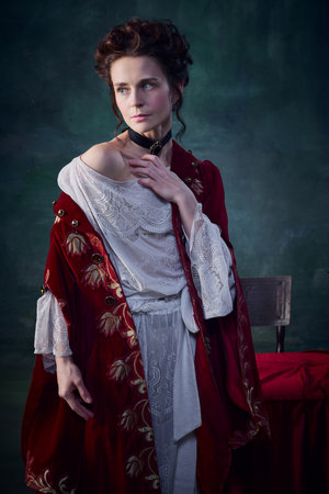 Portrait of attractive young woman with pale skin and blushing cheeks posing in velvet red dress...