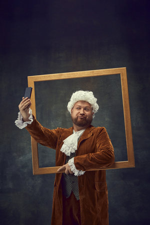 Posing. Vintage portrait of young man in brown vintage suit and white wig like medieval royal hunter isolated on dark background.の写真素材