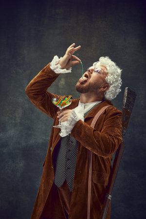 Eating jelly candies. Portrait of young man in brown vintage suit and white wig like medieval royal...