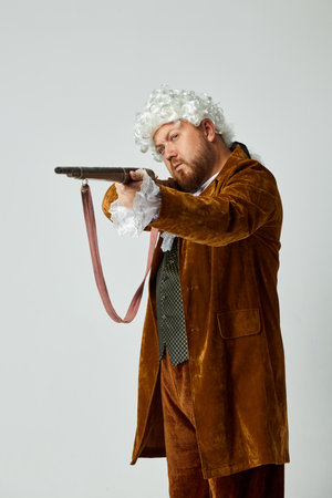 A fine shot. Young man in image of medieval person in vintage brown hunting suit and white wig with old hunting rifle isolated over grey background.の写真素材