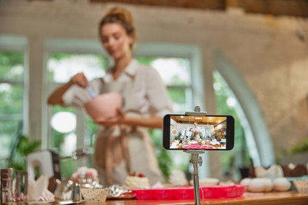 View through phone screen. Young beautiful girl culinary specialist writes video blog on baking. Smiling female vlogger or shooting blogの写真素材