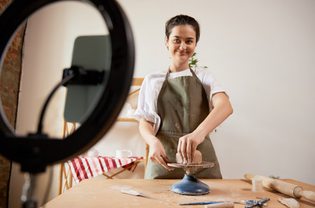 Charming smiling girl, pottery master with supplies recording vlog at home studio. Hobbies and leisure activities. Remote training, online classesの写真素材