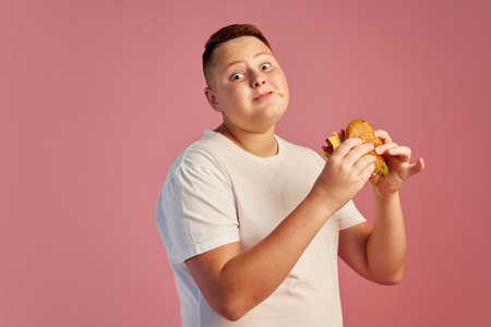 Half-length portrait of cute overweight boy in white t-shirt tasting delicious burger isolated on pink background. Fast food, taste, body positive, emotionsの写真素材