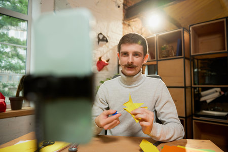 Young man sitting at home, holds a master class on origami and recording vlog at home studio....