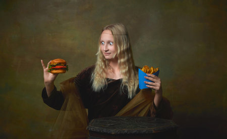 Cute young charming girl in image of Mona Lisa looking at hamburger with shock isolated on dark green background. Art, beauty, style, imitationの写真素材
