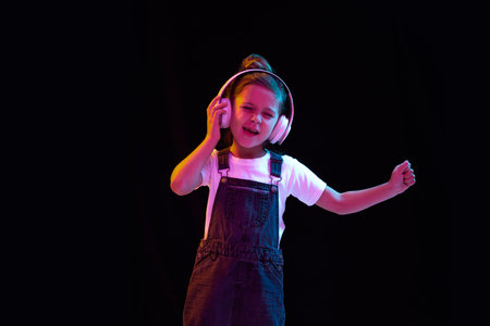 Music. Stylish little girl, pupil in white t-shirt and denim dress posing isolated over dark background in neon light. Concept of beauty, kids fashion, children emotionsの写真素材