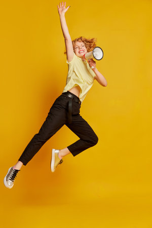 Full-length portrait of active young red-haired girl jumping with megaphone isolated on yellow studio background. Human emotions, facial expression, beauty concept.の写真素材