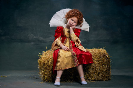 Portrait of little red-headed girl, child in costume of royal person sitting on hay with boring face isolated over dark green backgroundの写真素材