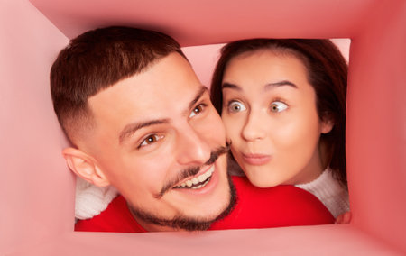 Portrait of lovely young couple, cheerful man and woman looking inside paper box isolated over pink background. Presentsの写真素材