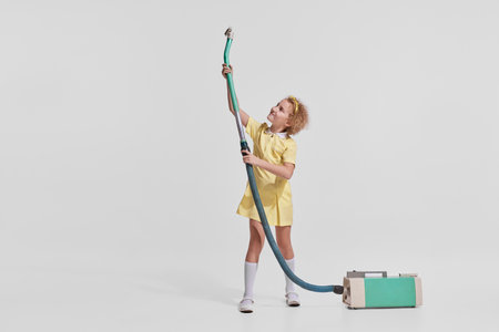Portrait of cute little girl, child in yellow dress cleaning with vacuum cleaner isolated over grey backgroundの写真素材