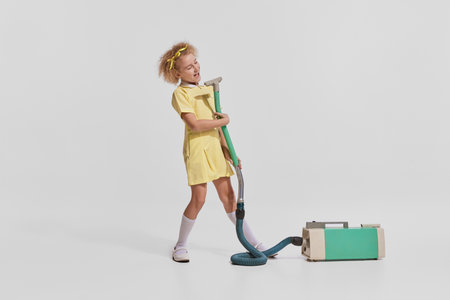 Portrait of cute little girl, child in yellow dress cleaning with vacuum cleaner and singing isolated over grey backgroundの写真素材