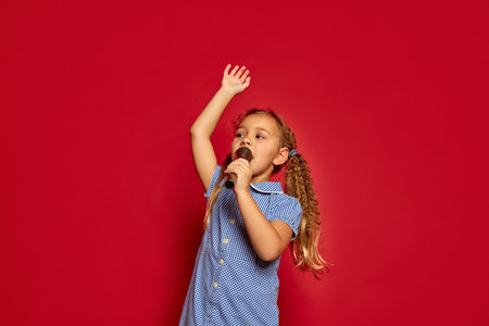 Cute little kid girl isolated on red studio background. Human emotions, facial expression. Concept of childhood, children, lifestyle. Copy space for ad. She listen music, sing song in microphoneの写真素材