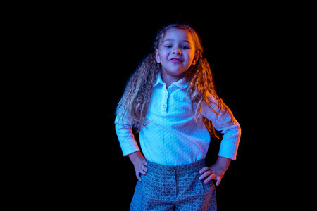 Charming little girl looking at camera isolated over dark background in neon light. Kids emotions,...