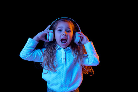 Music. One beautiful little girl, kid listening music in headphones isolated on blue color background in neon light. Human emotions, facial expression. Concept of childhood, children, beautyの写真素材