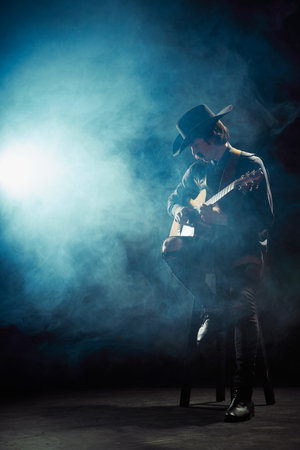 Portrait of man with moustaches in country style clothes playing guitar isolated over dark blue background with smoke. Jazz performerの写真素材
