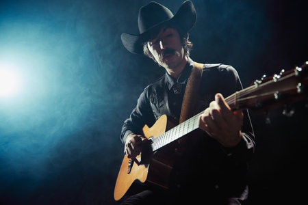 Portrait of man with moustaches in country style clothes playing guitar isolated over dark blue background with smokeの写真素材