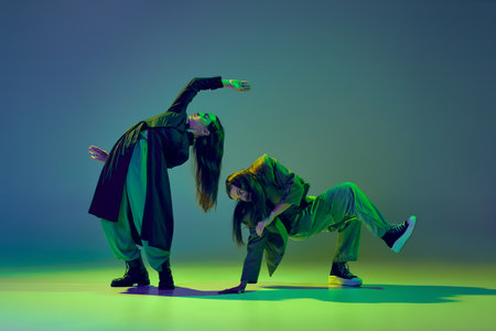 Inspiration. Young stylish flexible girls in modern attires dancing contemp style dance isolated over gradient blue-green background at dance hall in neon light.の写真素材