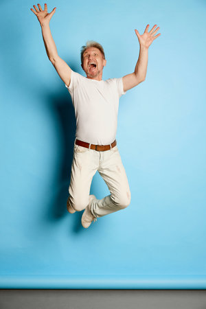 Portrait of mature man in white casual clothes posing, cheerfully jumping isolated over blue studio...