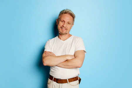 Portrait of mature man in white T-shirt posing isolated over blue studio background. Delightful and...