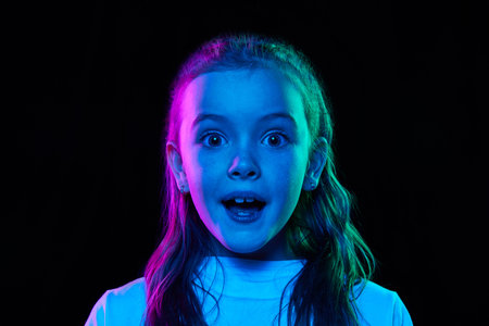 Portrait of little beautiful girl, child in white T-shirt posing with positive shock isolated over black studio background in neon lightの写真素材
