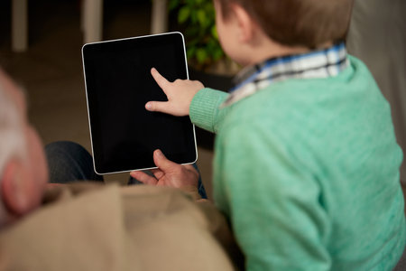 Back view of grandfather and grandson spending time together, using tablet at home. Concept of...