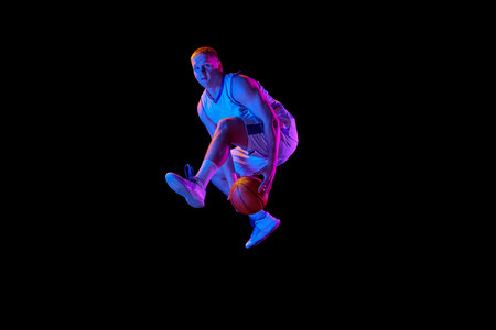 Dynamic portrait of young active athlete, male basketball player in sports uniform practicing with ball, jumping, dribbling isolated over dark background in neon lightの写真素材