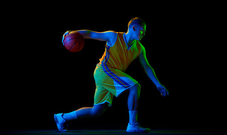 Studio shot of young active athlete, male basketball player in sports uniform in motion and action with ball isolated over dark background in neon lightの写真素材