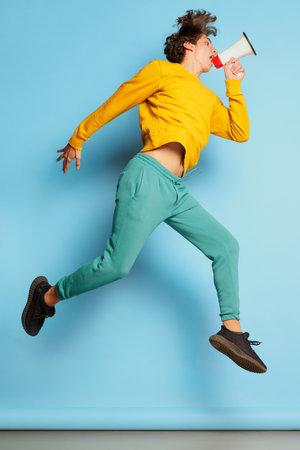 Portrait of young man with curly hair posing, jumping and talking in megaphone isolated over blue...