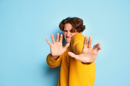 Portrait of young man with curly hair posing with hands forward, expressing fear isolated over blue backgroundの写真素材
