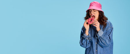 Portrait of young emotive girl in pink panama, posing, funny eating donut isolated over blue background. Flyerの写真素材