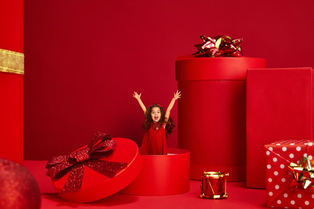 Happy cute little girl, child in beautiful dress appearing from present box over red background. Merry holidays. Giftsの写真素材