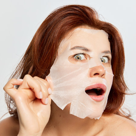 Portrait of beautiful, red-haired woman taking off moisturizing and revitalizing mask isolated on white background. Anti-wrinkles careの写真素材