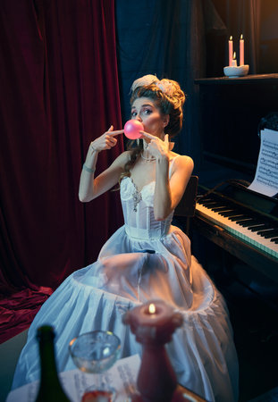 Portrait of young beautiful girl in image of medieval person in elegant white dress sitting at the piano and posing with bubblegum. Comparison of eras, beauty, history, artの写真素材