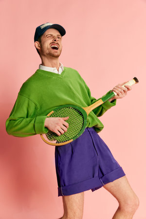 Portrait of young man in vintage sportive clothes playing tennis racket like guitar isolated over pink background.の写真素材