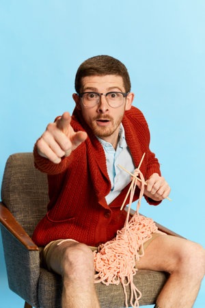 Portrait of young man sitting on chair, knitting isolated over blue background. Attention lookの写真素材