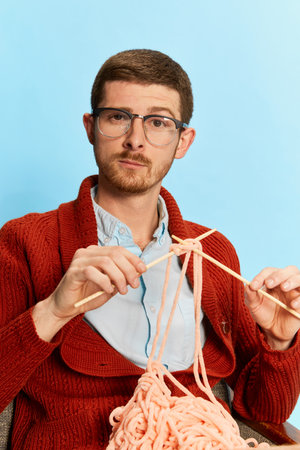 Portrait of young man sitting on chair and knitting isolated over blue background. Evening relaxationの写真素材