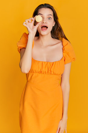 Portrait of beautiful brunette girl in cute dress posing with sweet dessert, macaron over orange backgroundの写真素材