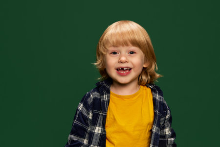 Portrait of cute little boy, child in yellow T-shirt and checkered shirt posing with smile over green studio background. Concept of childhood, emotionsの写真素材