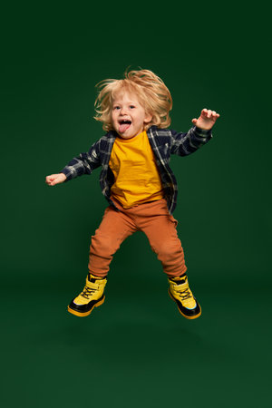 Full-length portrait of cheerful little boy, child posing, jumping, having fun over green studio background. Concept of childhood, emotionsの写真素材