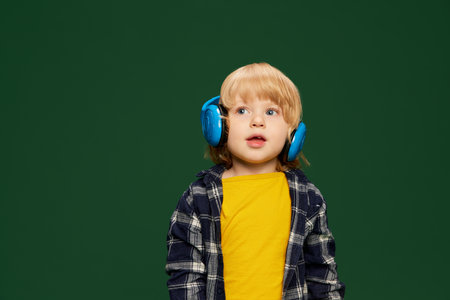 Portrait of little boy, child posing in headphones, listening to fairy tales over green studio background. Concept of childhood, emotionsの写真素材