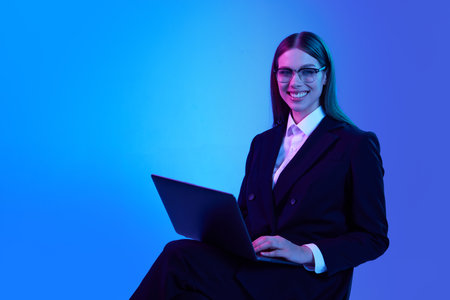 Woman in stylish official suit working on laptop over blue background in neon light. Successful business womanの写真素材