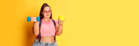 Portrait of overweight young woman posing with dumbbell and apple over yellow studio background. Diet and sport. Bannerの写真素材