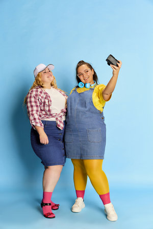 Portrait of two overweight women, friends taking photo, selfie with camera, posing over blue studio...