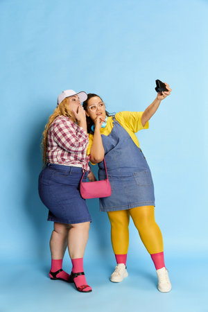 Portrait of two women, friends taking photo with camera, posing over blue studio background. Cheerful meetingの写真素材