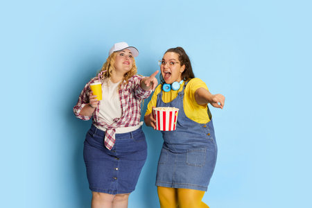 Portrait of two fat, beautiful women, friends posing with popcorn basket over blue studio...