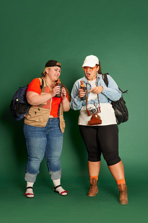 Portrait of two overweight women, friends in casual clothes with backpacks, eating hot dogs over...