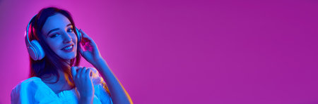 Portrait of young girl in white blouse, listening to music in headphones over purple background in...
