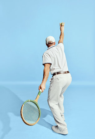 Back view. Serving. Portrait of handsome senior man in stylish white outfit playing tennis over blue background. Concept of leisure activity, hobby, lifestyleの写真素材