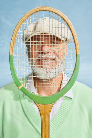 Portrait of handsome senior man in stylish white outfit posing, looking through tennis racket over sky blue background. Concept of leisure activity, hobby, lifestyleの写真素材