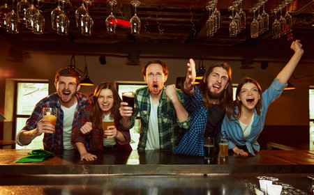 Group of young people, men and women watching match at pub and drinking beer. Fans emotionally...